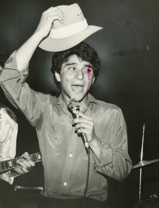 A young man in a shiny shirt sings into a microphone while raising a hat over his head on darke…