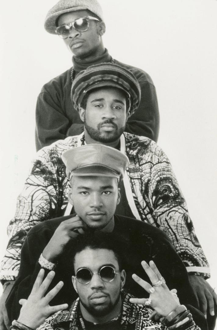 Four black men stacked on top of each other wearing colourful clothes and caps in front of whit…