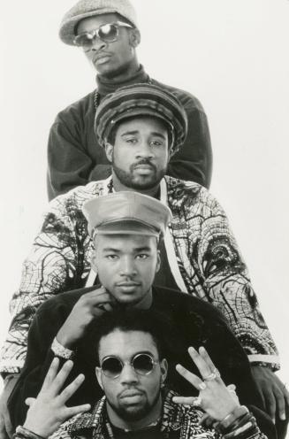 Four black men stacked on top of each other wearing colourful clothes and caps in front of whit…