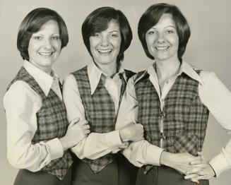 Three identical young women wearing plaid vests and white shirts hold each other's arms.