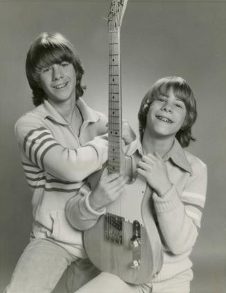 Two boys, brothers with similar sweaters, jeans and shaggy blond haircuts cradle an electric gu…