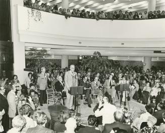 Picture of a man at a microphone speaking to a seated crowd at library opening. 