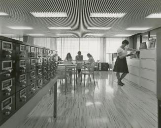 Picture of interior of a library showing card catalogue, staff looking in at something from a f…