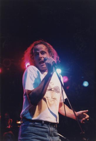 Middle aged man in t-shirt and jeans sings into microphone on darkened stage.