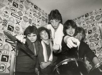 five teen boys crowd behind a drum kit in the corner of a domestic room. two of the boys point …