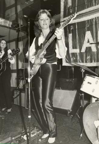 On darkened stage, full body image, young woman with light coloured curly hair plays an electri…