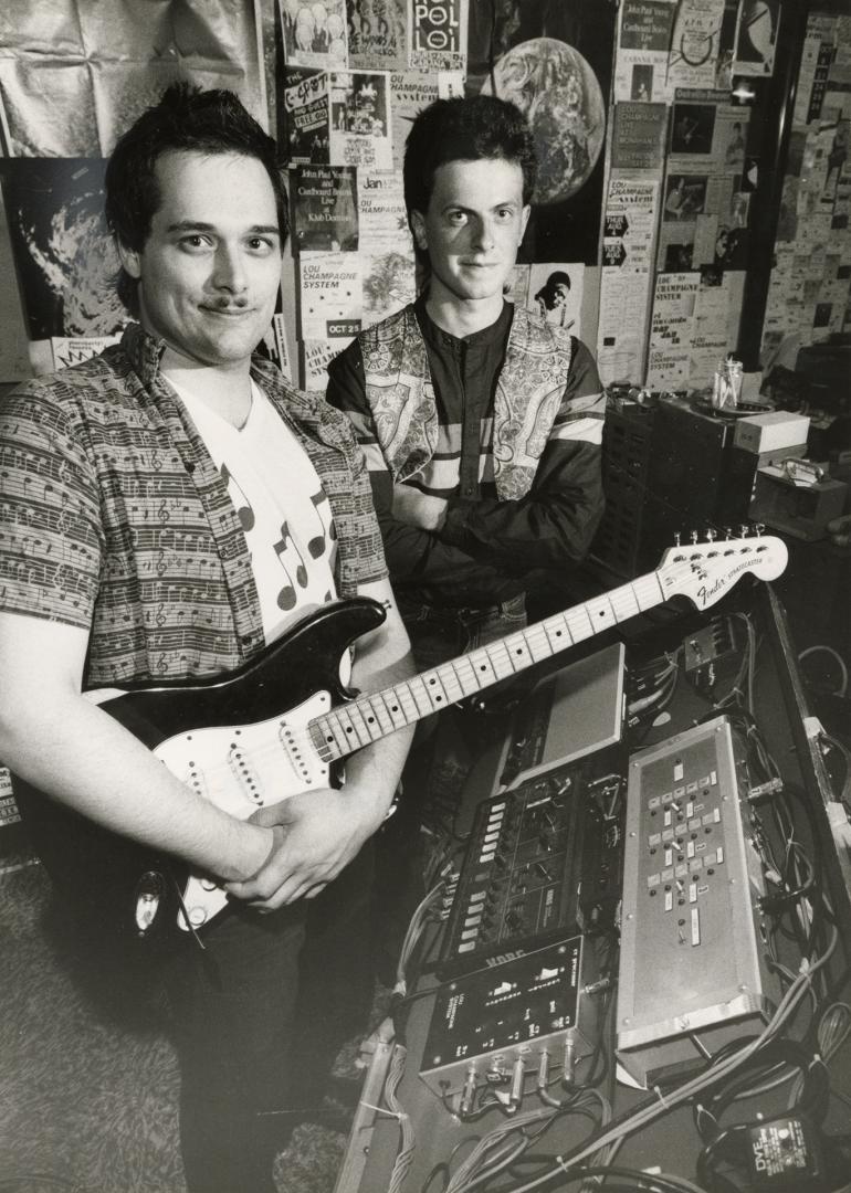 Beside console in recording studio, man with electric guitar and behind on his right another ma…
