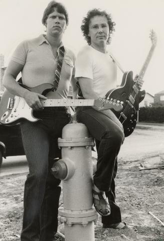 Two young men outside leaning on a fire hydrant carrying electric guitars. 