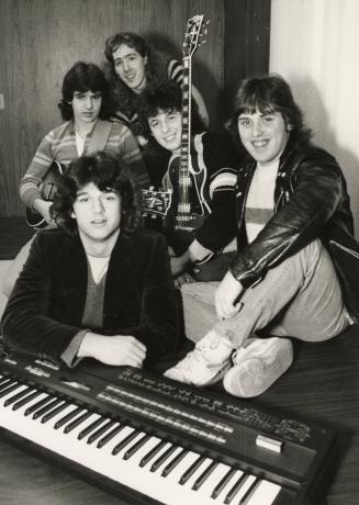 Five young men dressed in jeans with shaggy haircuts in a tight circle behind a keyboard in a w…