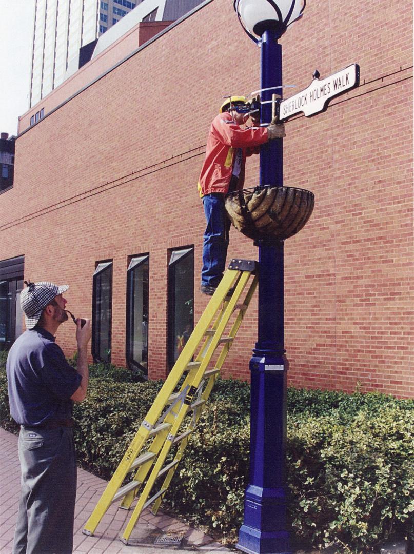 Colour image of man on ladder leaning against pole, attaching a sign beside a brick building. A…