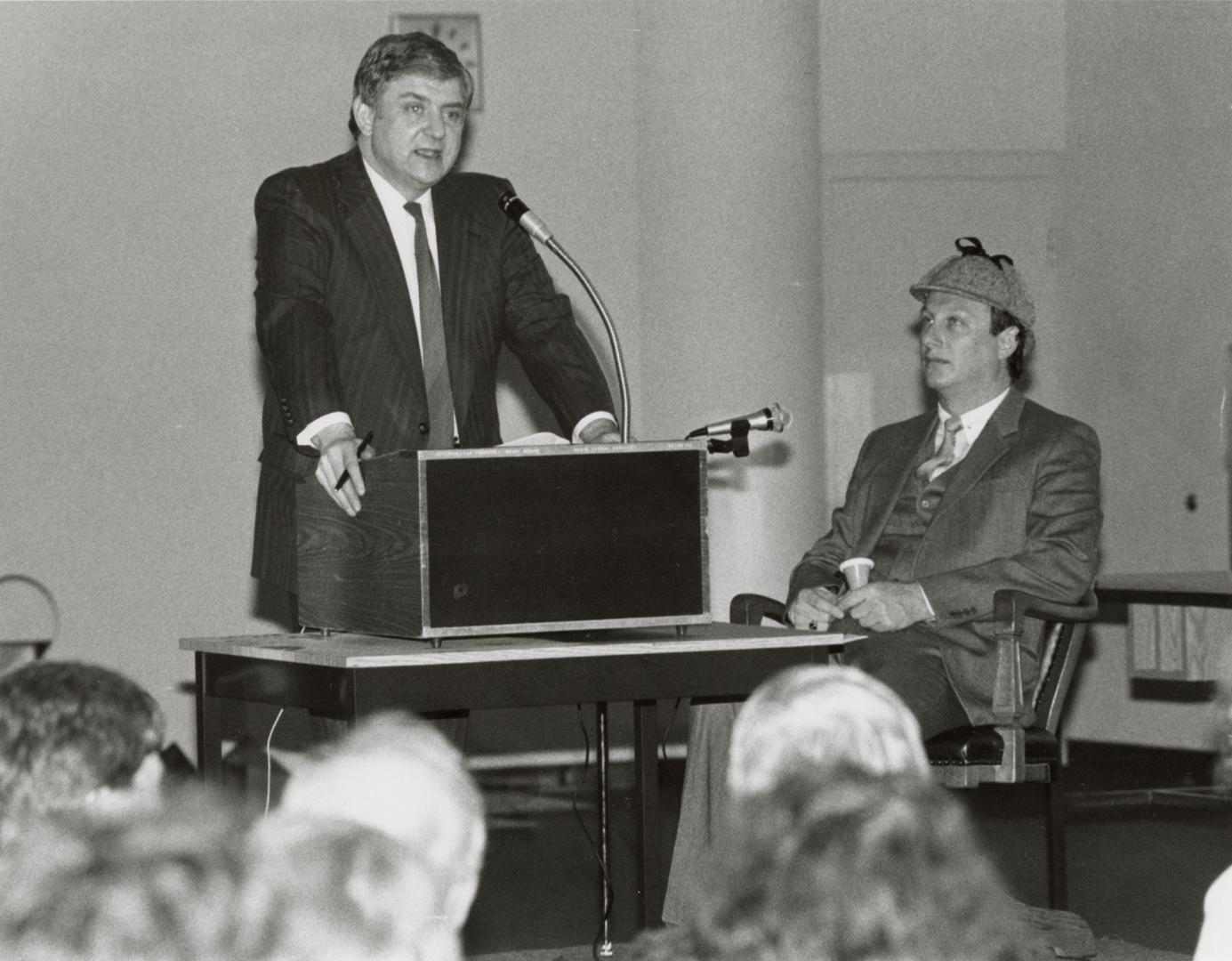 Man standing at podium giving a lecture to the left, to the right, man sits in Sherlock Holmes …