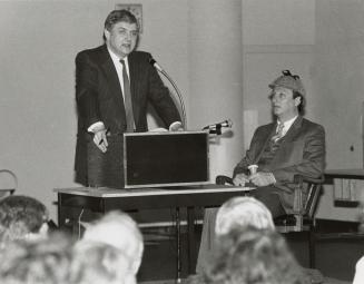 Man standing at podium giving a lecture to the left, to the right, man sits in Sherlock Holmes …