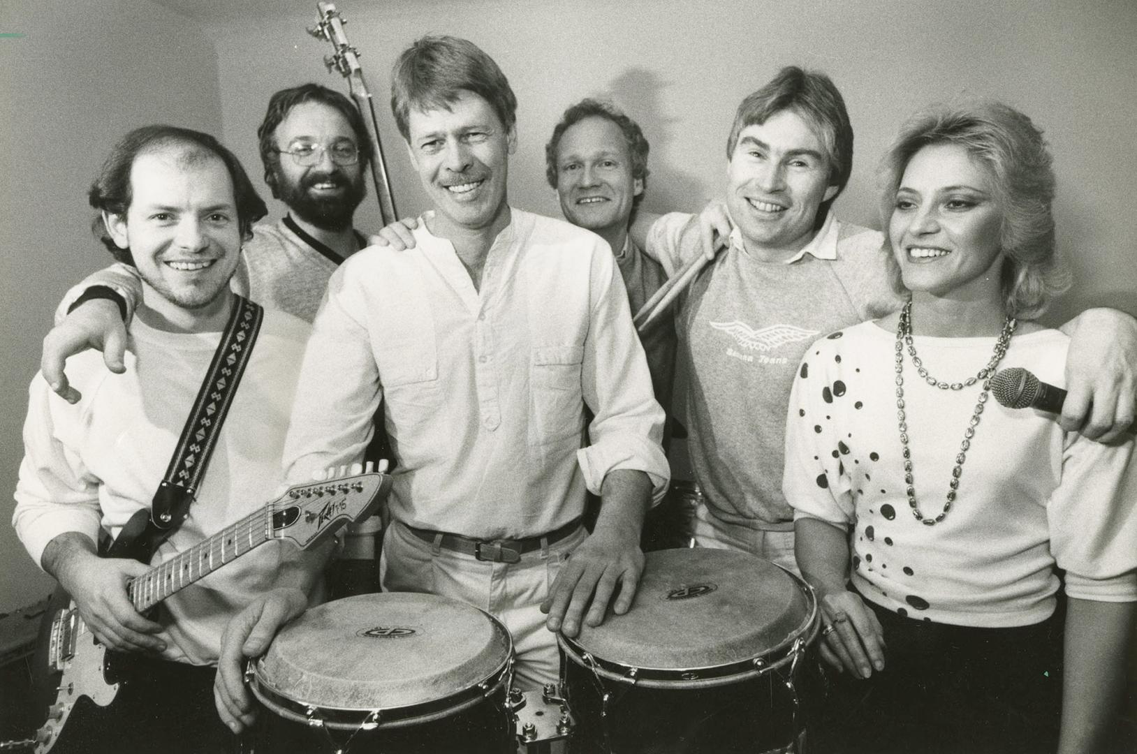 five men and a woman on the far right in a row with guitars and bongos. 