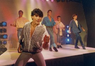 A group of kid and teen boys dance and sing on a stage wearing jeans and large shirts. 