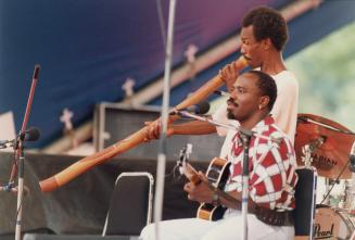 two middle aged men, foreground sits one playing guitar, behind him the other stands playing a …