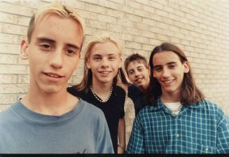 Four teen boys in front of a brick wall with longish hair, all smiling. 