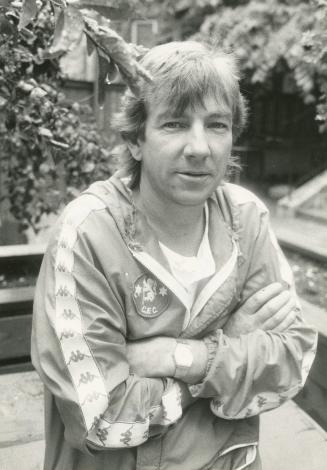 Middle aged man surrounded by foliage, his arms crossed, wearing a sporty jacket and a metal wa…
