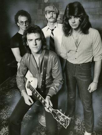 Young man sitting with guitar on his knee surrounded by three young men standing. All wearing j…