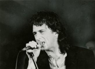 Man with dark long curly hair wearing open neck and dark blazer sings into hand-held microphone…