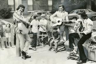 Daytime on street corner, three men in sport coats and boots playing guitars and singing with a…