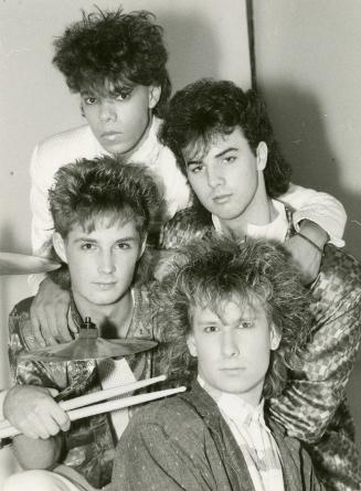 Four young men in a circular pattern with big coiffed hair in fancy jackets. The one on bottom …