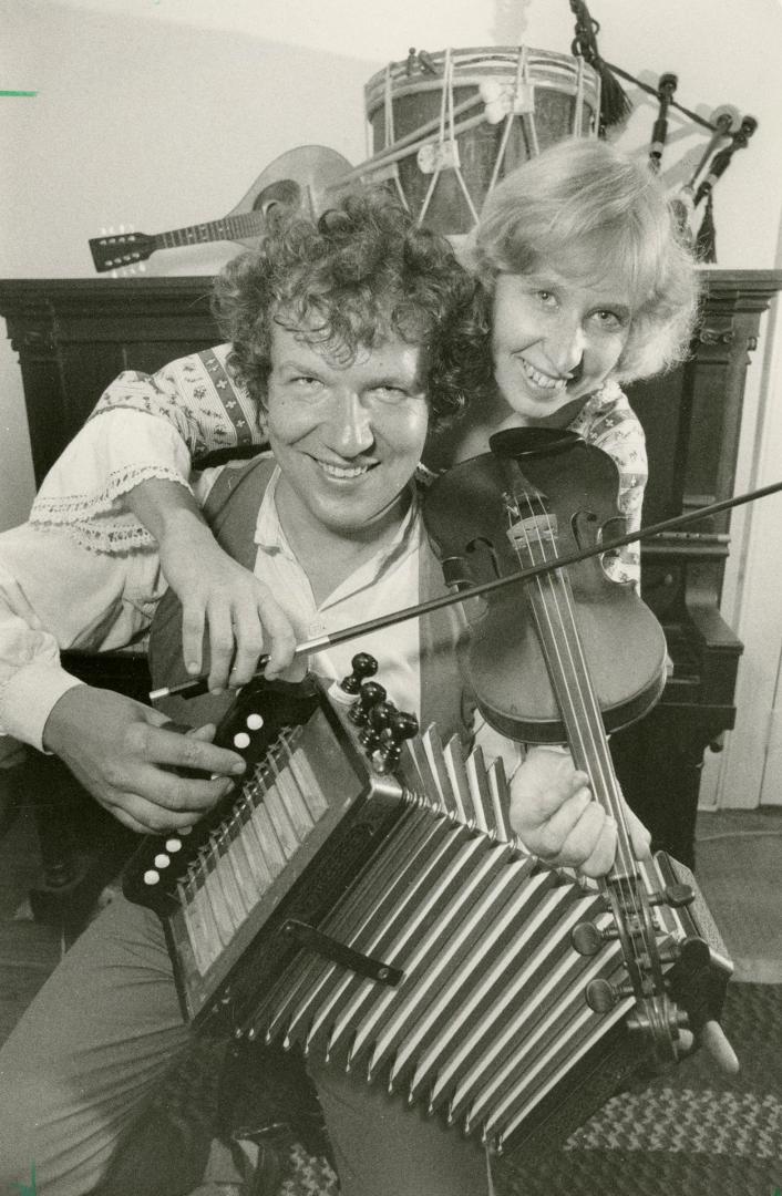 A man sitting with an accordion in his hands is hugged by a woman behind him holding a violin. …