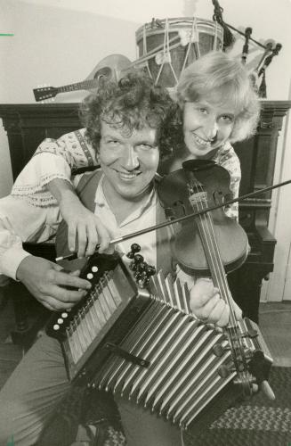 A man sitting with an accordion in his hands is hugged by a woman behind him holding a violin. …