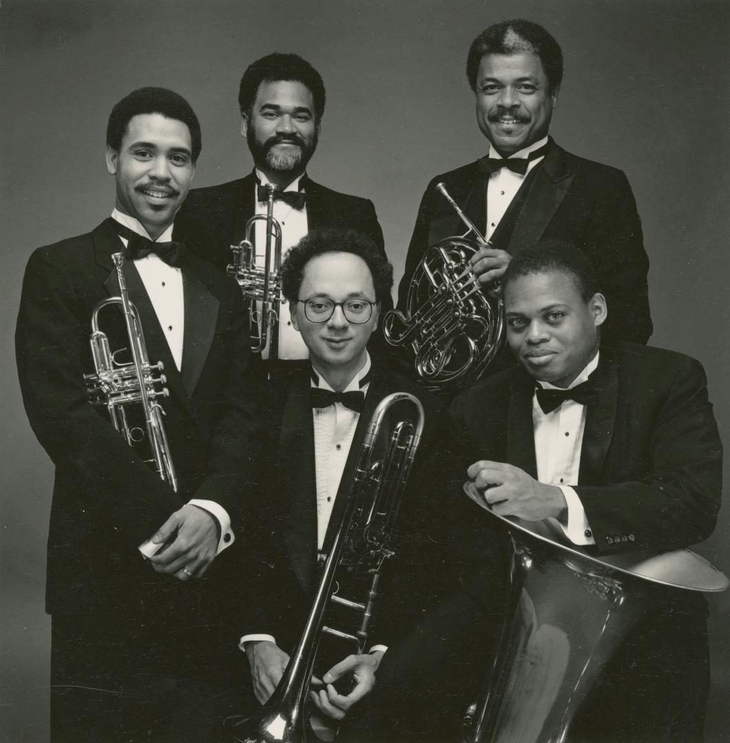 Five men in tuxedos and bow ties in a circular group, each holding a different brass instrument…
