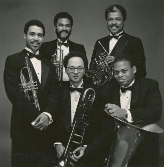 Five men in tuxedos and bow ties in a circular group, each holding a different brass instrument…