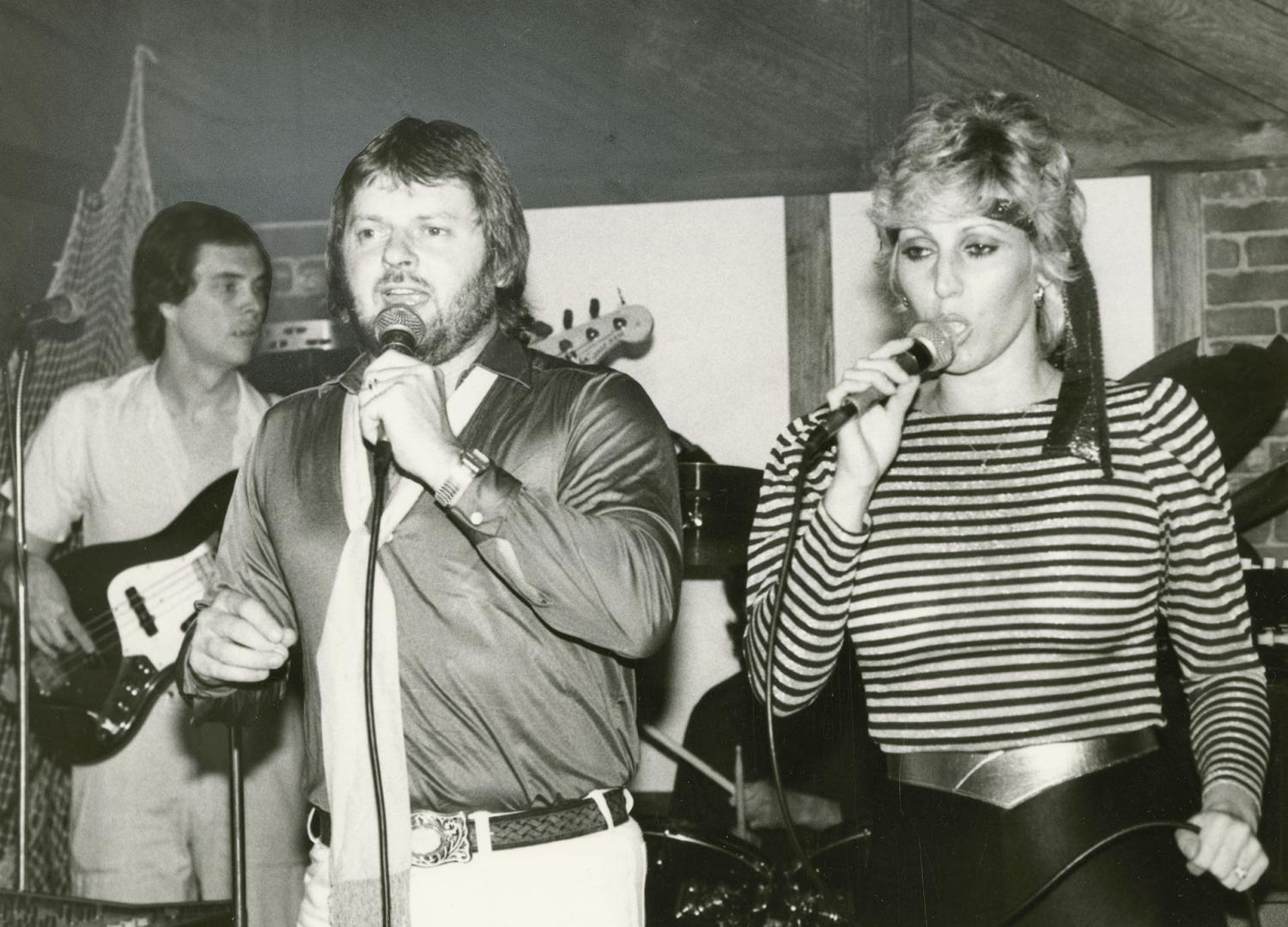 A man and a woman in a low ceilinged room sing into handheld microphones, a guitarist behind th…