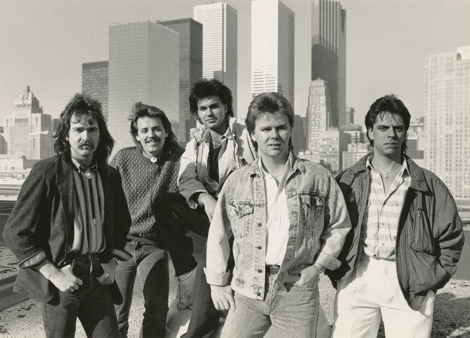 Five young men with shaggy hair in jeans on rooftop with downtown skyline behind them. Daytime,…