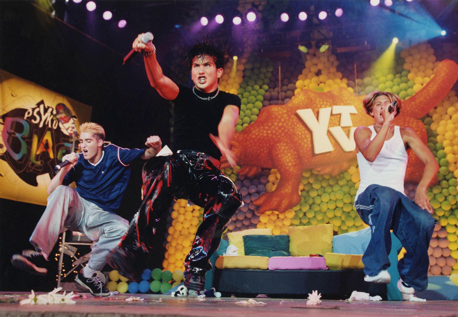Three young men jumping in crouch position with hand-held microphones on stage in front of colo…