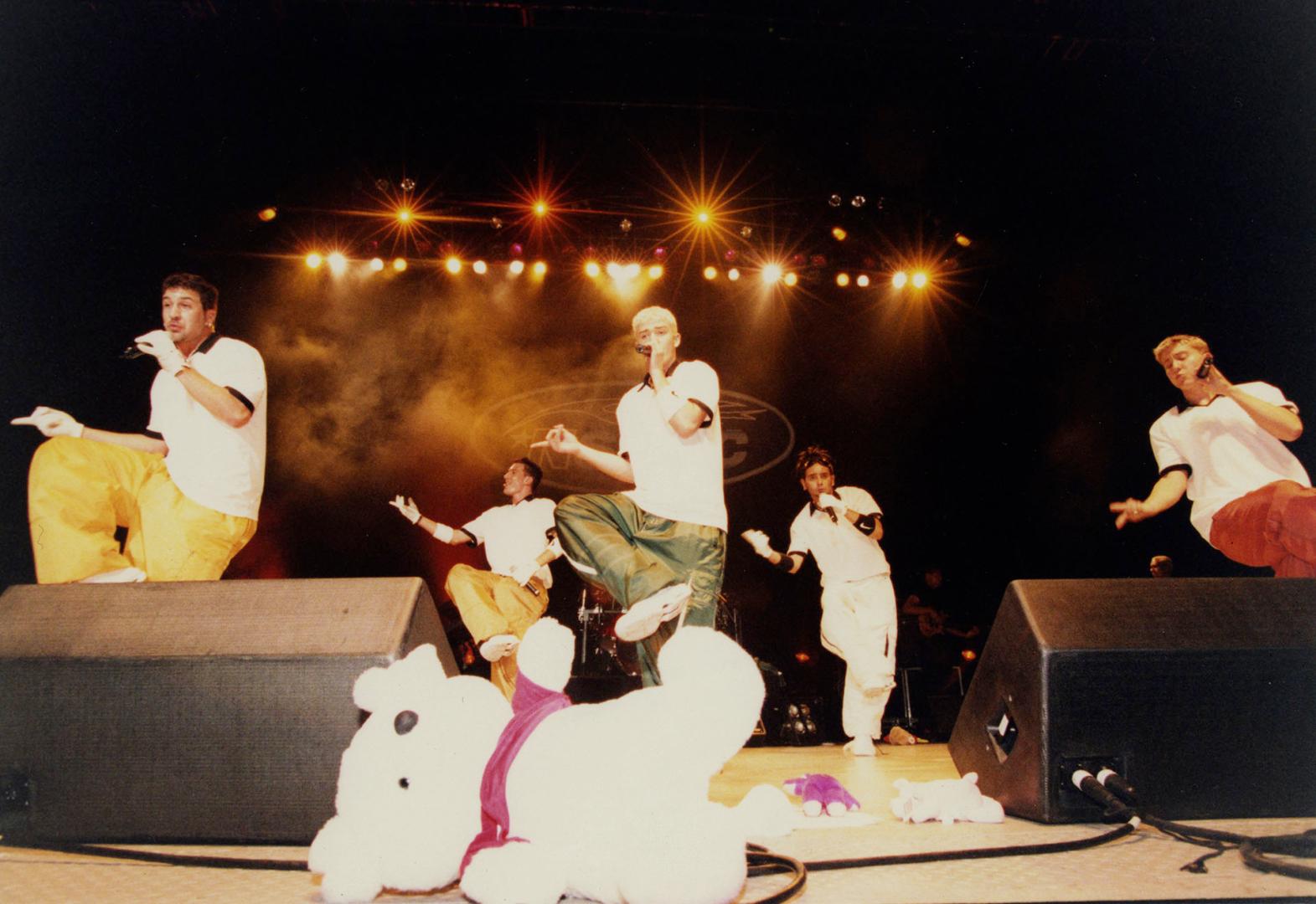 Five young men dancing with one knee up behind stage monitor speakers. Lights overhead. 