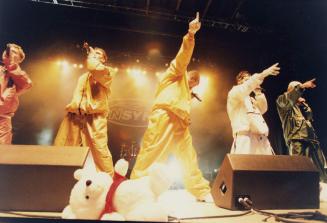 Five young men in loose clothing pointing up into the air behind monitor speakers, a big stuffe…