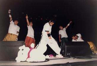 Five young men in white t-shirts and baggy pants on stage behind monitor speakers and a large w…