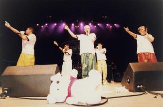 Five young men in white t-shirts and baggy pants, all pointing their arms up to the left, three…