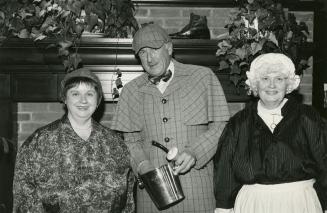 Black and white photograph of two women and a man standing in front of a fire place. Woman on t…