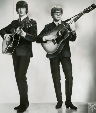 Two young men, full body shot from floor, in suits, shirts and ties and heeled boots, carrying …
