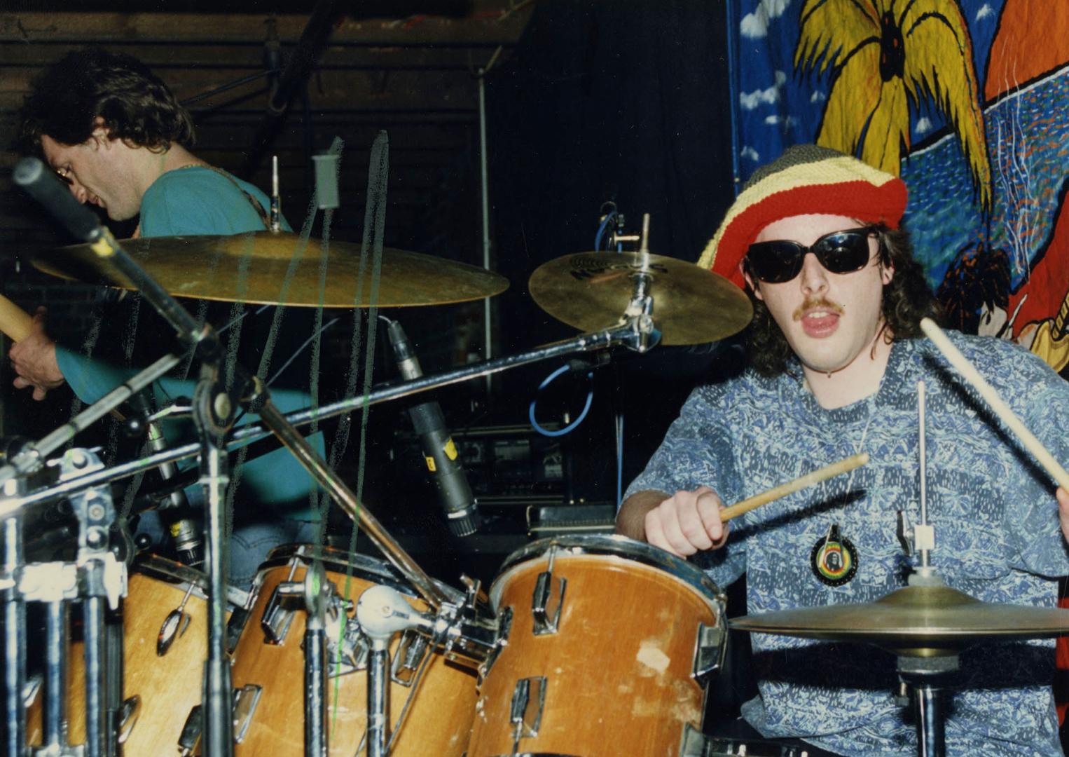 Young man in sunglasses and green/yellow/red beret playing drumkit on darkened stage with guita…