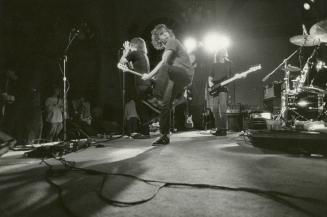 On darkened stage, Two young men with guitars, one kicks his leg up. In front of them, another …
