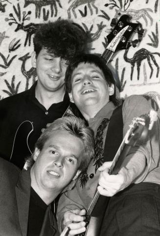Three young men, two brunette and one blond, in a tight grouping, wearing shirts, two hold elec…