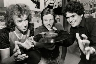 Three young men intently watch a vinyl record spinning on the middle person's thumb in a record…