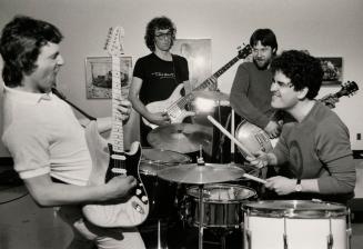 Three young men playing guitars crowd around the drumkit and drummer wearing t-shirts and jeans…
