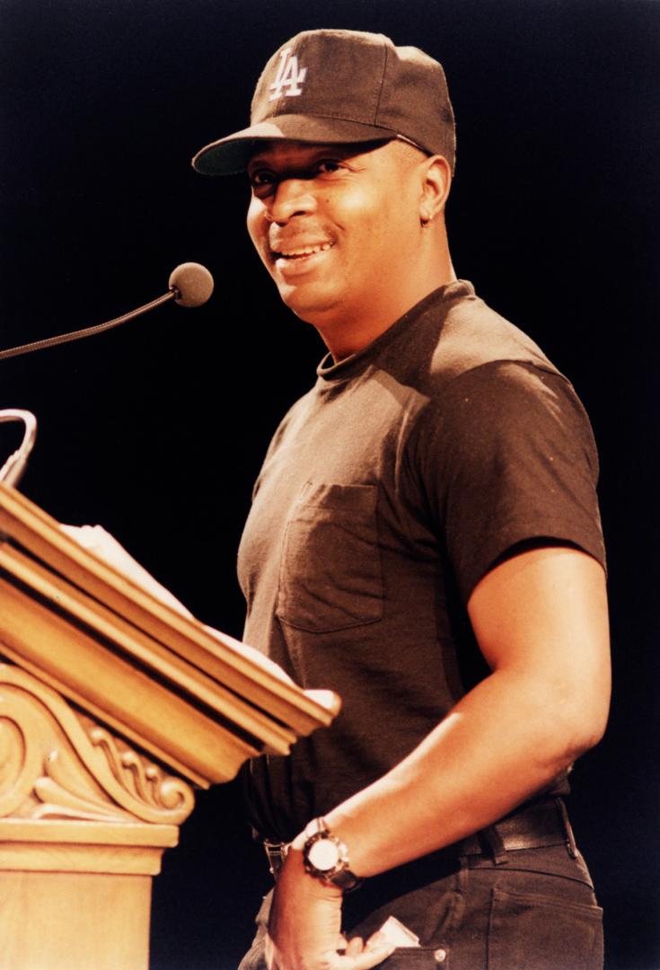 Man in ballcap and brown T-shirt and gold watch smiling behind lectern with microphone.