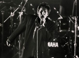 Young man in blazer and plaid shirt sings into microphone on darkened stage with drumkit behind…