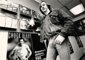 Young man in jean jacket and jeans and dark t-shirt stands beside a Chuck Berry poster and lean…