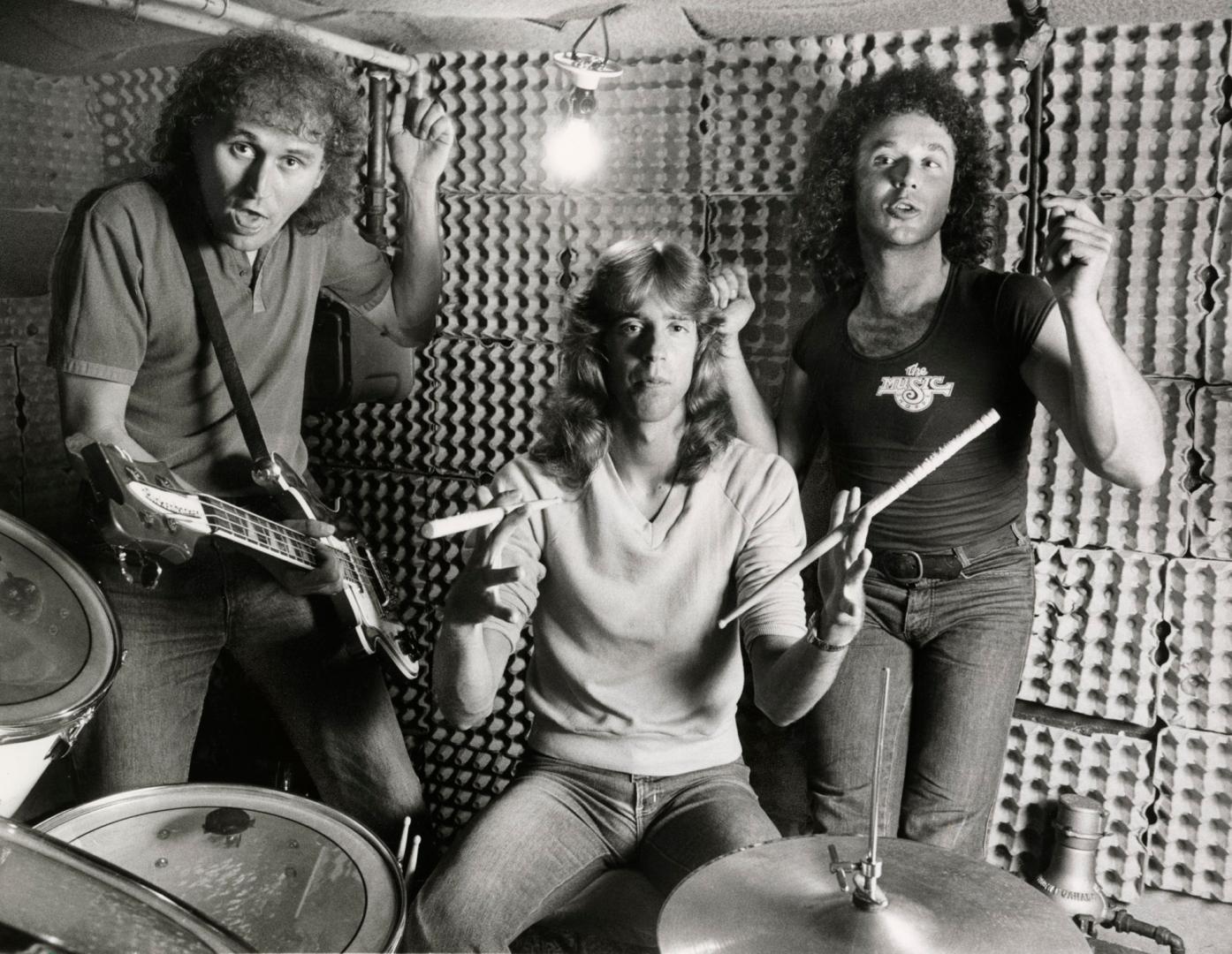 Three young men in egg carton-lined practice space, behind drummer and drumkit. All wear tight …