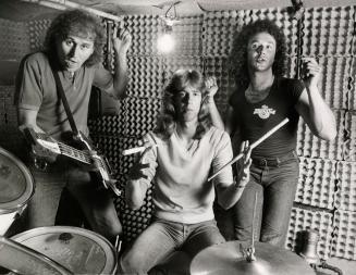 Three young men in egg carton-lined practice space, behind drummer and drumkit. All wear tight …