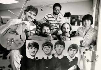Posed behind a large poster of the moptop era Beatles are four men. The one on the left holds a…
