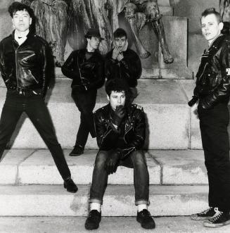 5 young men in black jeans and black leather motorcycle jackets lounge on the steps of a large …